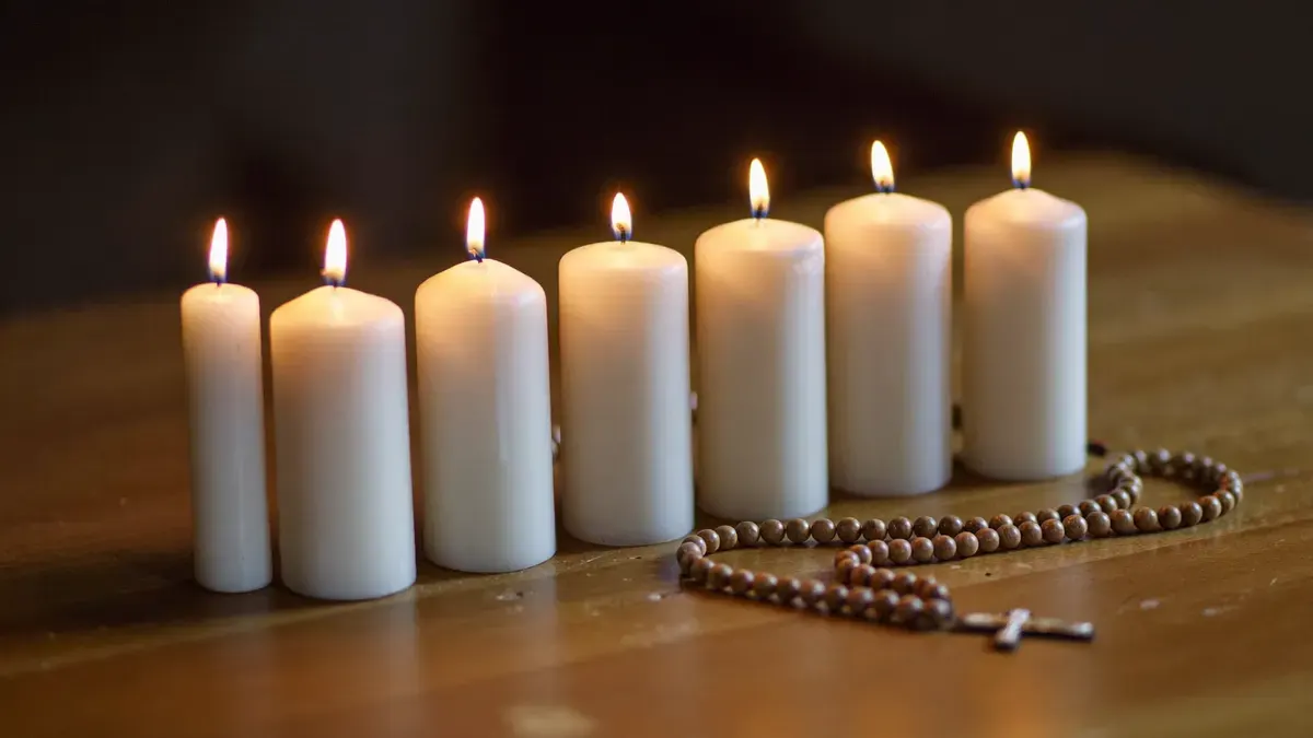 Velas brancas acesas enfileiradas sobre uma mesa de madeira, com um terço ao lado