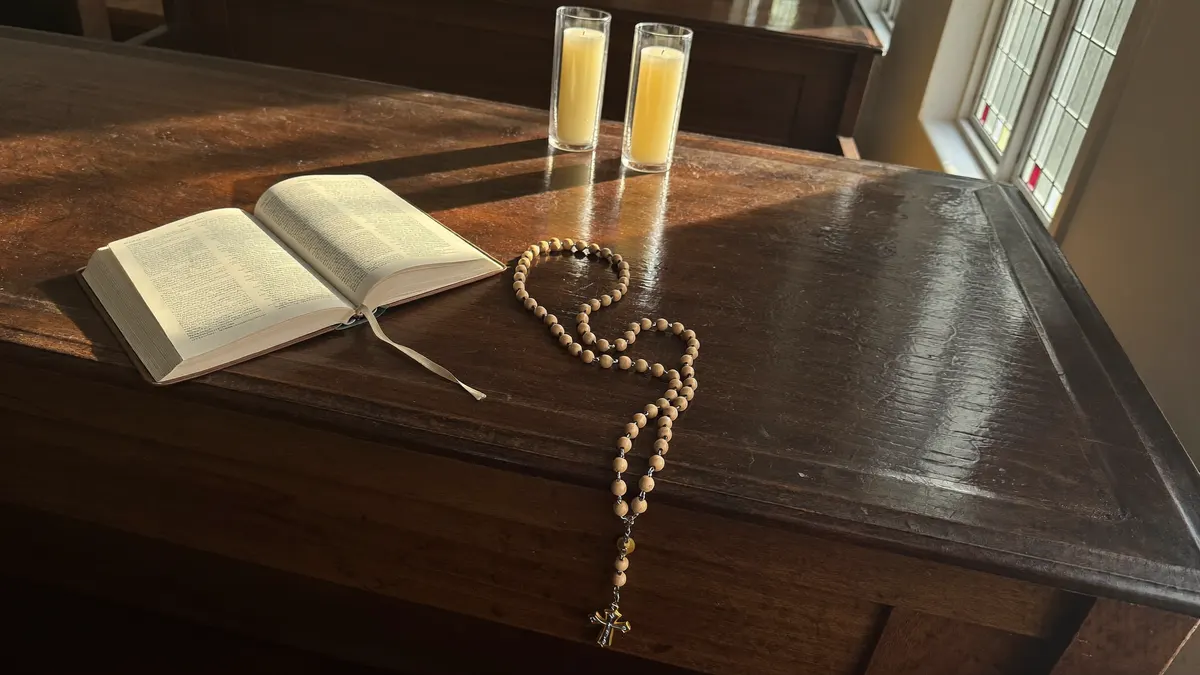 Rosário, Bíblia aberta e velas acesas sobre um banco de igreja, iluminados por luz suave de vitral