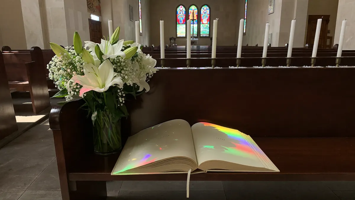 Velas acesas, flores brancas e um missal aberto sobre um banco de igreja, em ambiente silencioso e iluminado por luz suave