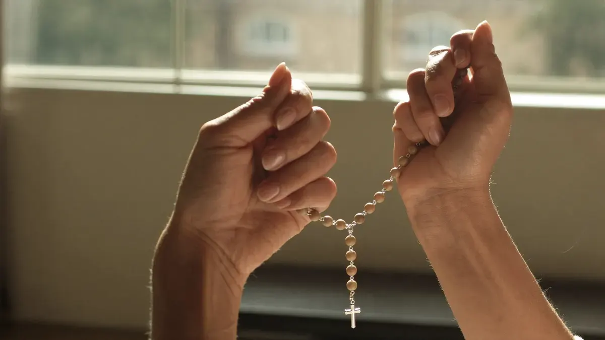 Mulher rezando o terço em silêncio perto de uma janela, mãos em destaque