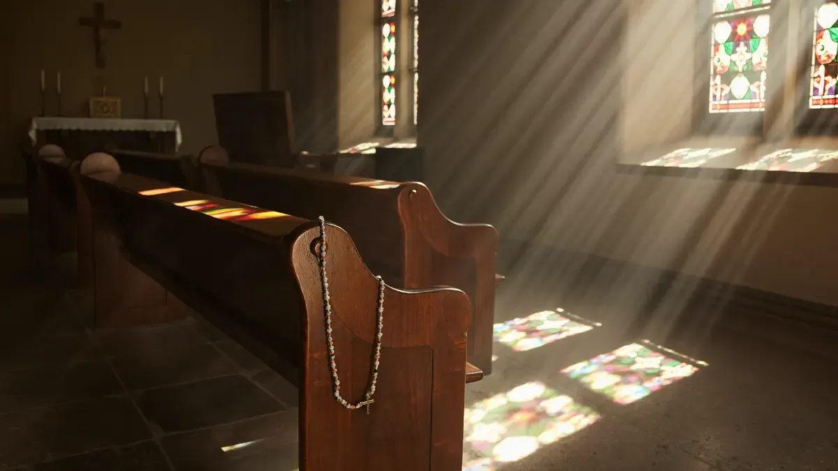 Luz suave entrando por um vitral em uma capela silenciosa, com um terço sobre o banco